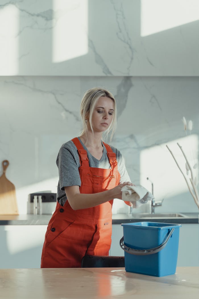 A woman wearing orange overalls cleans a kitchen counter with a rag and pail in a modern kitchen.